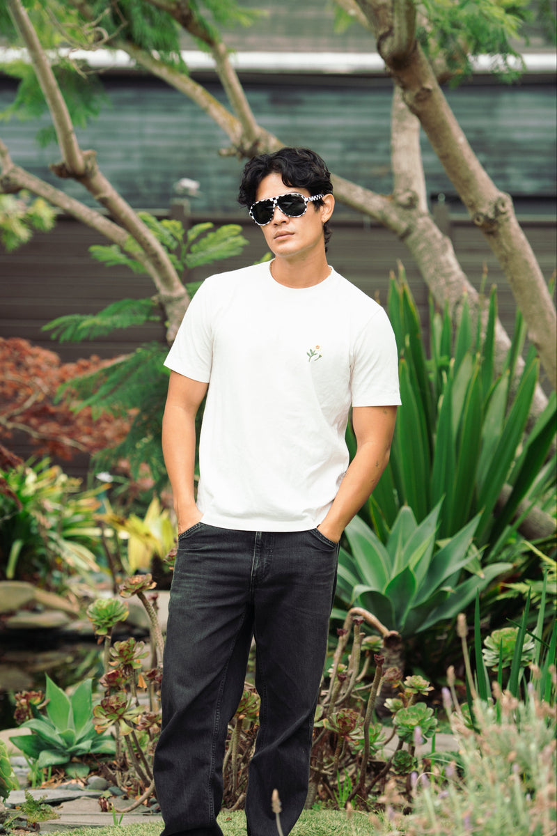 A man wearing a white crew-neck t-shirt with a small green chest graphic, black jeans, and sunglasses, standing in a lush green garden.